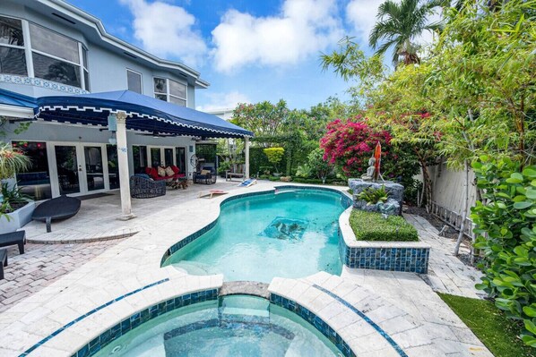 A swimming pool with a tiled deck, a covered seating area with chairs, and lush greenery. - Amazing house with pool (Miami)