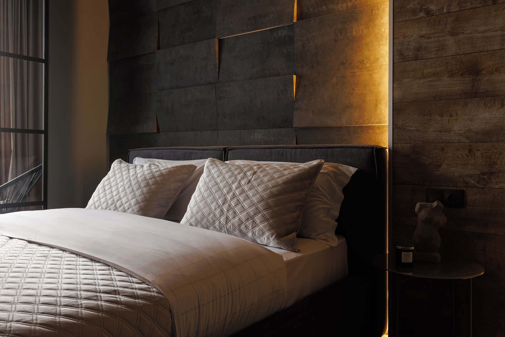 A modern bedroom with a dark headboard, quilted pillows, and a bedside table with a candle.