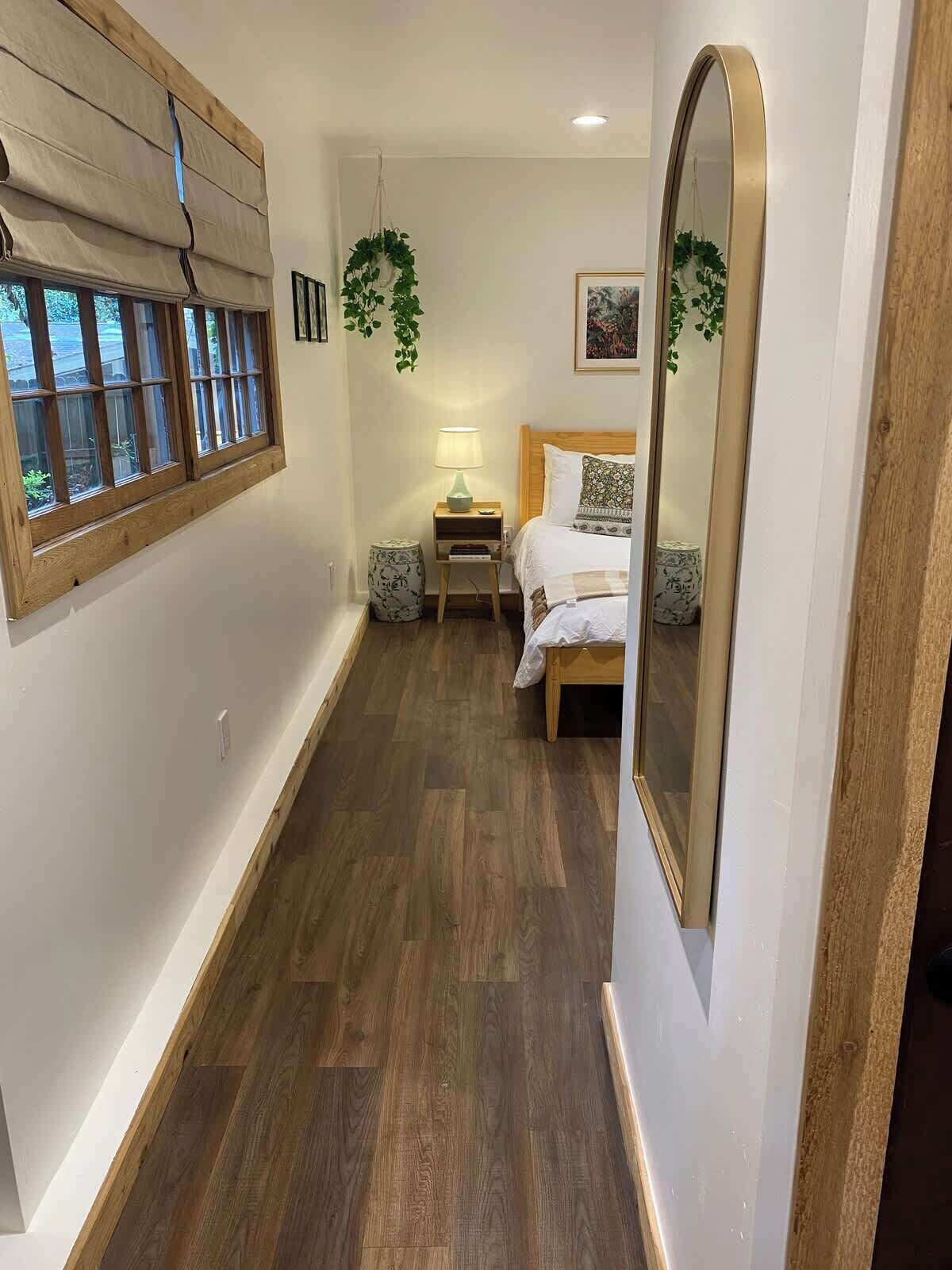 Habitación de hotel con piso de madera, cama con ropa blanca, mesita de noche con lámpara, planta colgante y un espejo grande.
