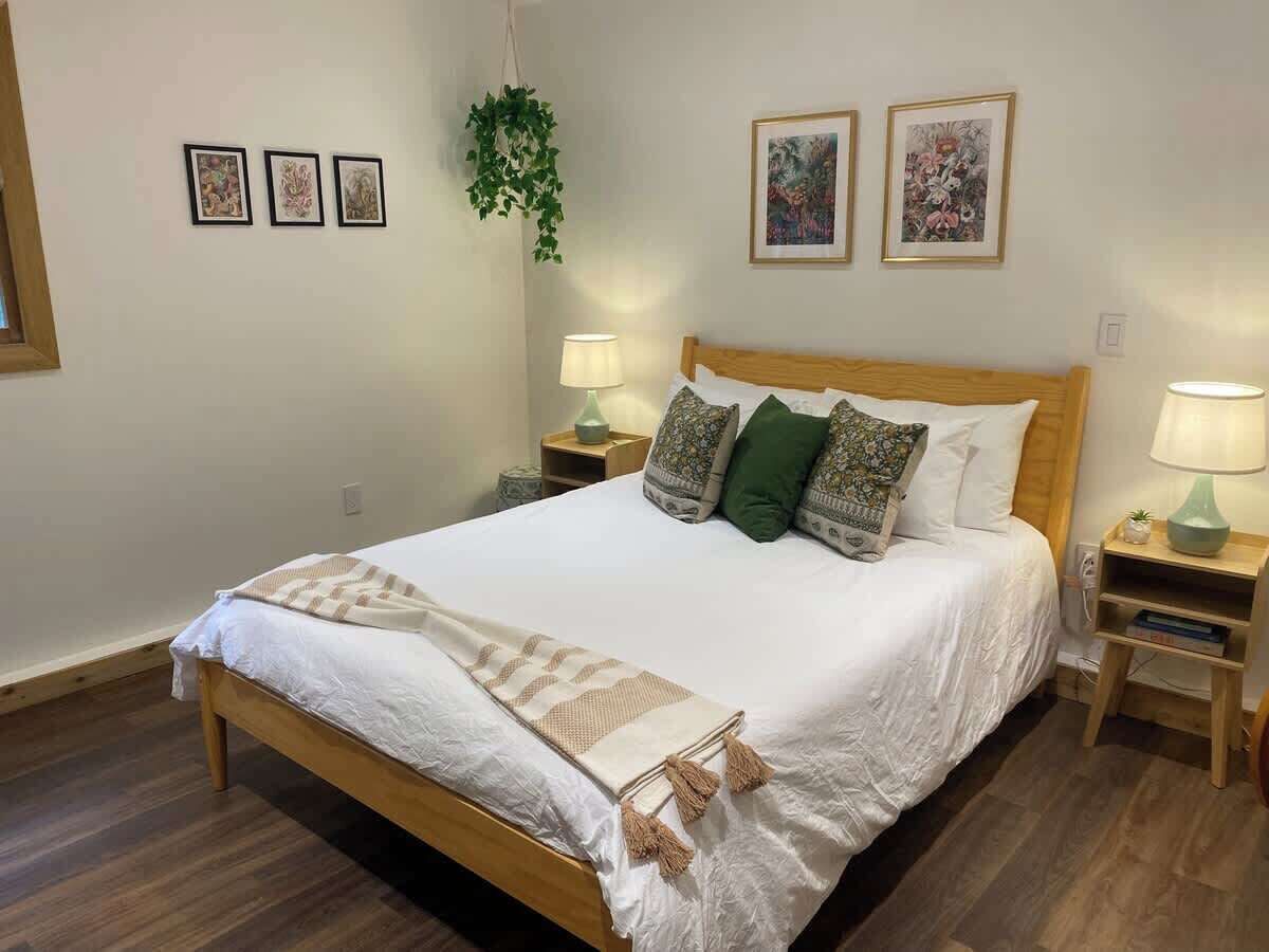 Un lit bien fait, recouvert d’une couette blanche et d’un plaid beige, flanqué de deux tables de chevet avec des lampes, d’une plante suspendue et de trois œuvres d’art encadrées accrochées au mur.
