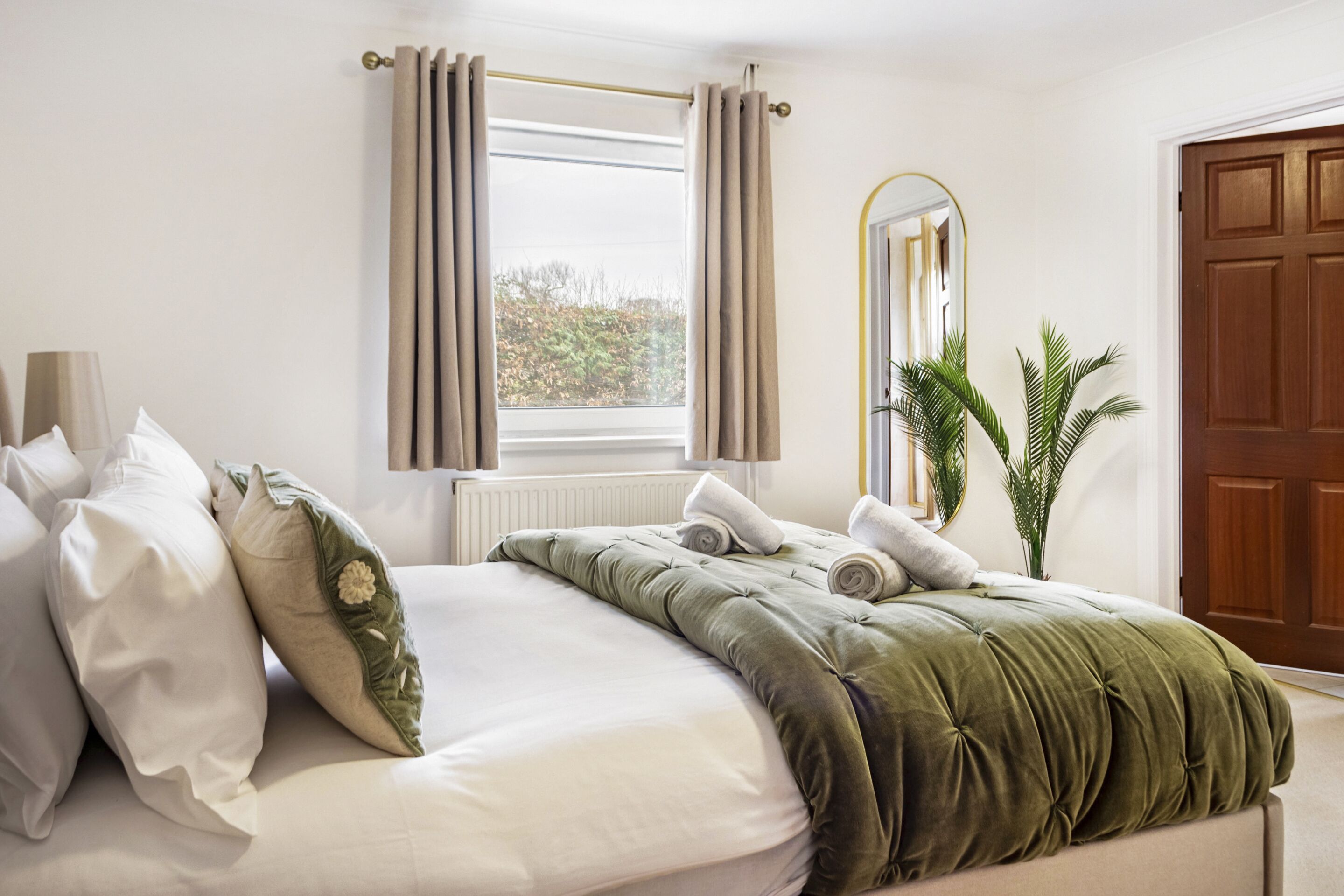 Ein Schlafzimmer mit Bett, Kissen, einem Fenster mit Vorhängen, einem Heizkörper, einem Spiegel, einer Tür und einer Pflanze.