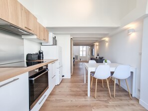 A modern kitchen with white cabinets, a black countertop, and a wooden ceiling. There is a dining area with a white table and chairs. - [Élégant] Duplex Centre-Ville Accès privé Plage (Arcachon)