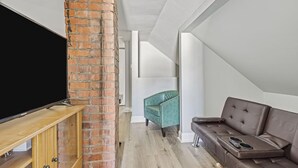 Ein modernes Wohnzimmer mit Flachbildfernseher auf Holzfuß, einer Ziegelwand, einem braunen Ledersofa und einem sesselartigen Sessel in Türkis.
