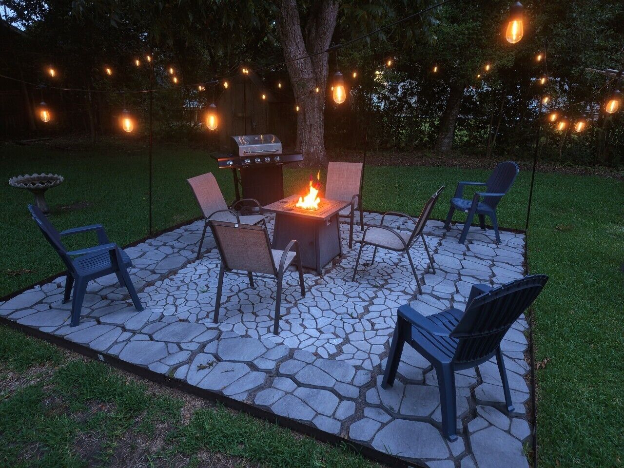 An outdoor patio with a stone fire pit, chairs, and a grill.