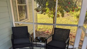 Ein überdachter Balkon mit zwei Korbstühlen und einem kleinen Tisch, der auf einen Garten mit Baum und Rasen blickt.