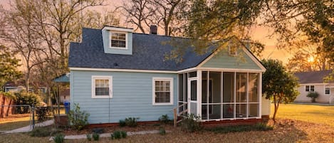 Einbungalow mit Veranda, einer kleinen angebauten Garage und einem Garten mit Bäumen und Rasen.