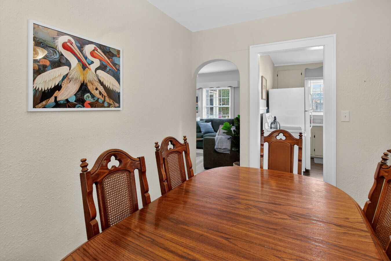 A dining area with a wooden table and chairs, a painting on the wall, and a view into a living room.
