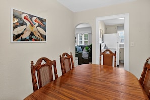 A dining area with a wooden table and chairs, a painting on the wall, and a view into a living room.
