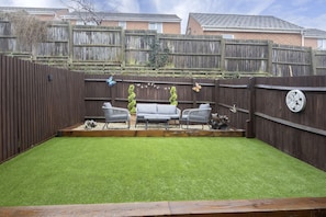Un jardin avec une pelouse artificielle, une clôture en bois, un canapé et deux chaises.