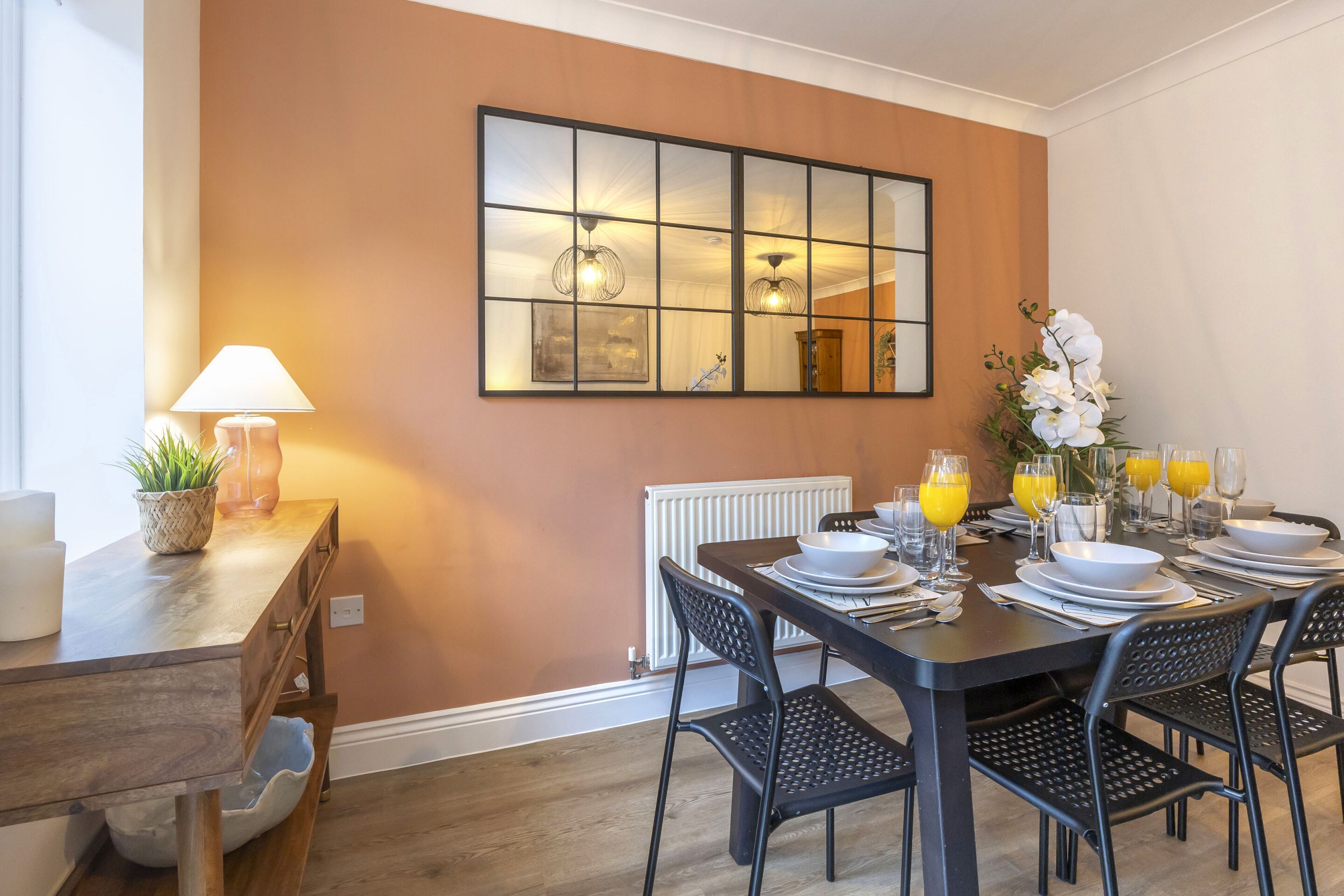 A dining area with a table set for four, a mirror on the wall, and a lamp on a side table.