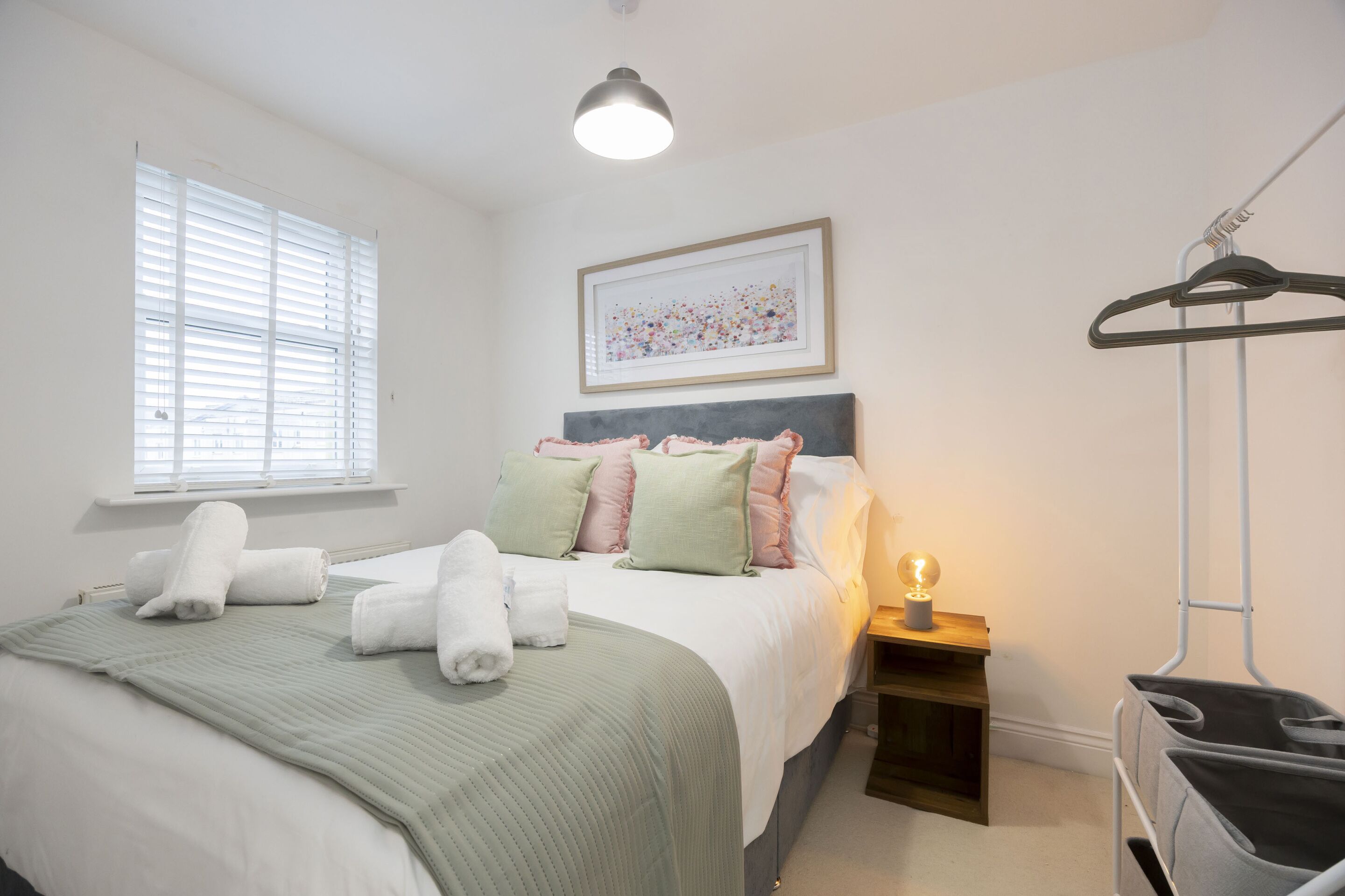 A neatly made bed with a green blanket, pillows, and rolled towels. There is a bedside table with a lamp and a framed picture on the wall.