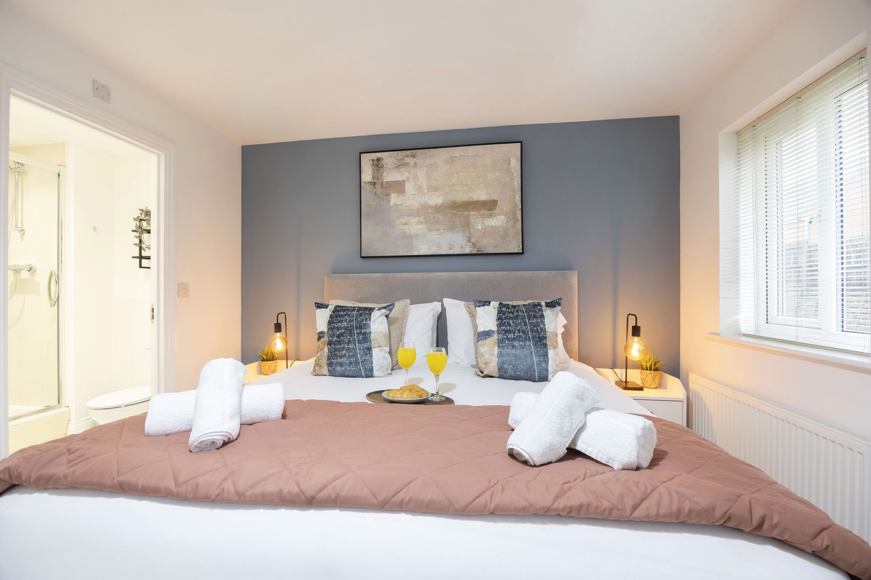 A neatly made bed with a brown bedspread, two white towels, and a plate with a croissant and two glasses of orange juice.