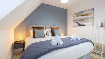 A modern bedroom with a large bed, a nightstand with a lamp, a painting on the wall, and a clothing rack.