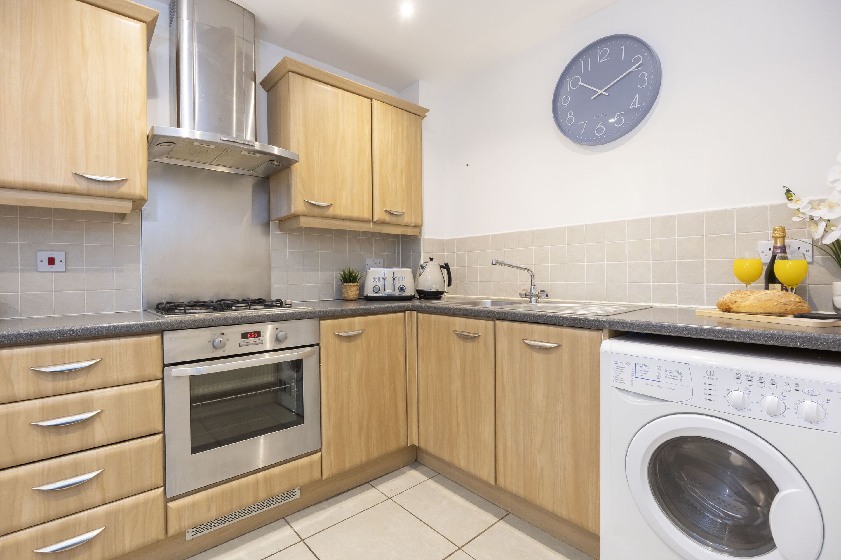 A modern kitchen with wooden cabinets, a built-in oven, a washing machine, and a clock on the wall.
