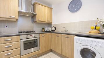 A modern kitchen with wooden cabinets, a built-in oven, a washing machine, and a clock on the wall.