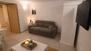 Una sala de estar con un sofá, un televisor montado en la pared, una mesita con una vela y una planta, y una puerta.
