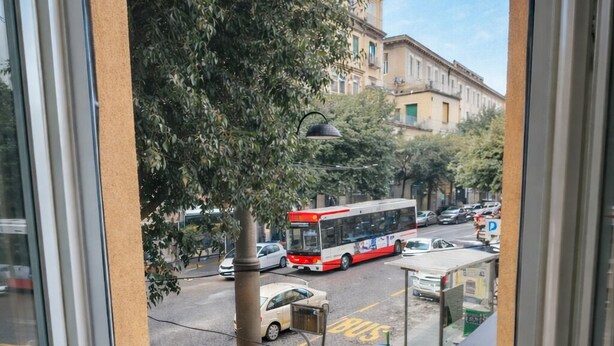 Una ventana con vistas a una calle, un autobús rojo y dos macetas con flores rojas.