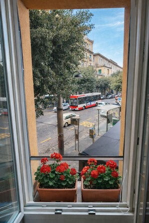 A window with a view of a street, a red bus, and two potted plants with red flowers. - St. Teresa Tower (Napoli)