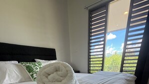Una cama bien hecha con sábanas blancas y una toalla enrollada encima, cabecera negra y vistas a la vegetación a través de las persianas.