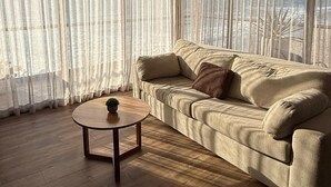 Una sala de estar con un sofá beige, una mesa de centro redonda de madera y cortinas translúcidas.