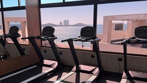 Un gimnasio con cintas de correr y equipamiento de ejercicio, amplias ventanas con vistas a un paisaje urbano.