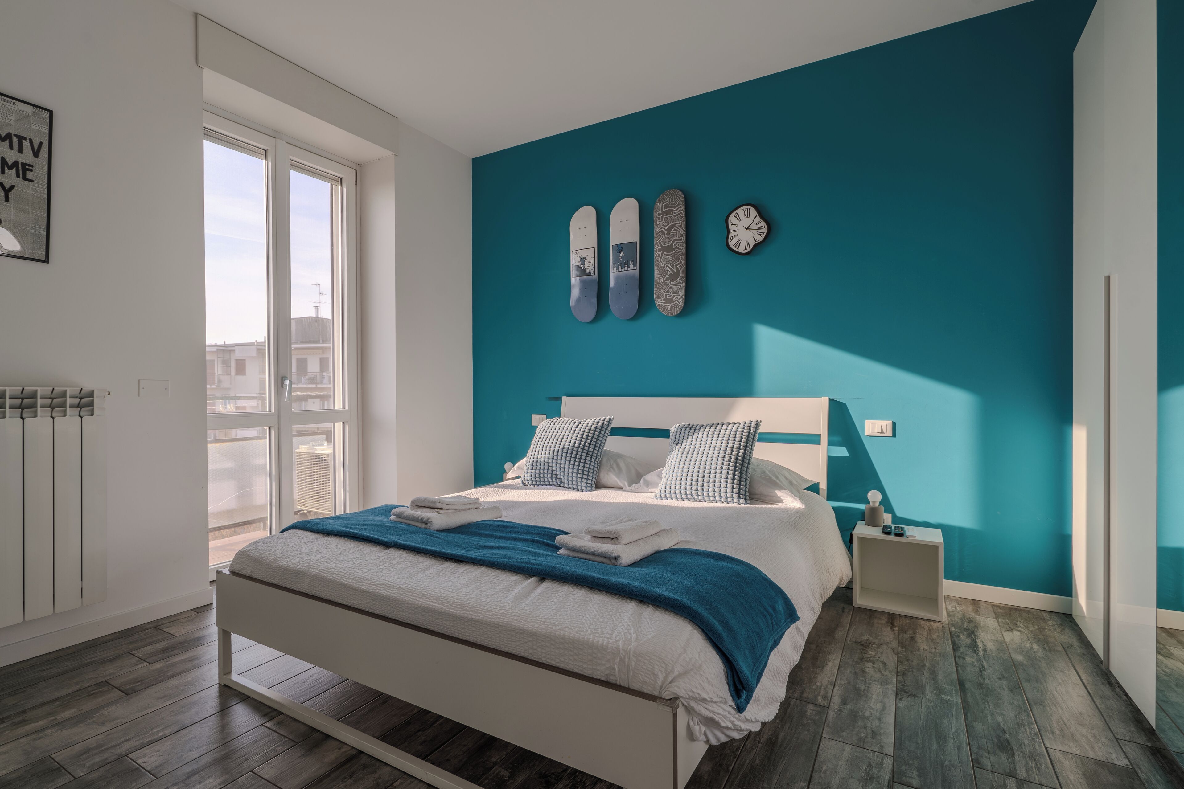 Un dormitorio moderno con cama, mesita de noche, ventana y una pared de acento color aguamarina.