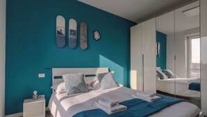Quarto moderno com paredes verde-água, uma cama grande com roupa de cama branca, um cobertor azul e três skateboards na parede.