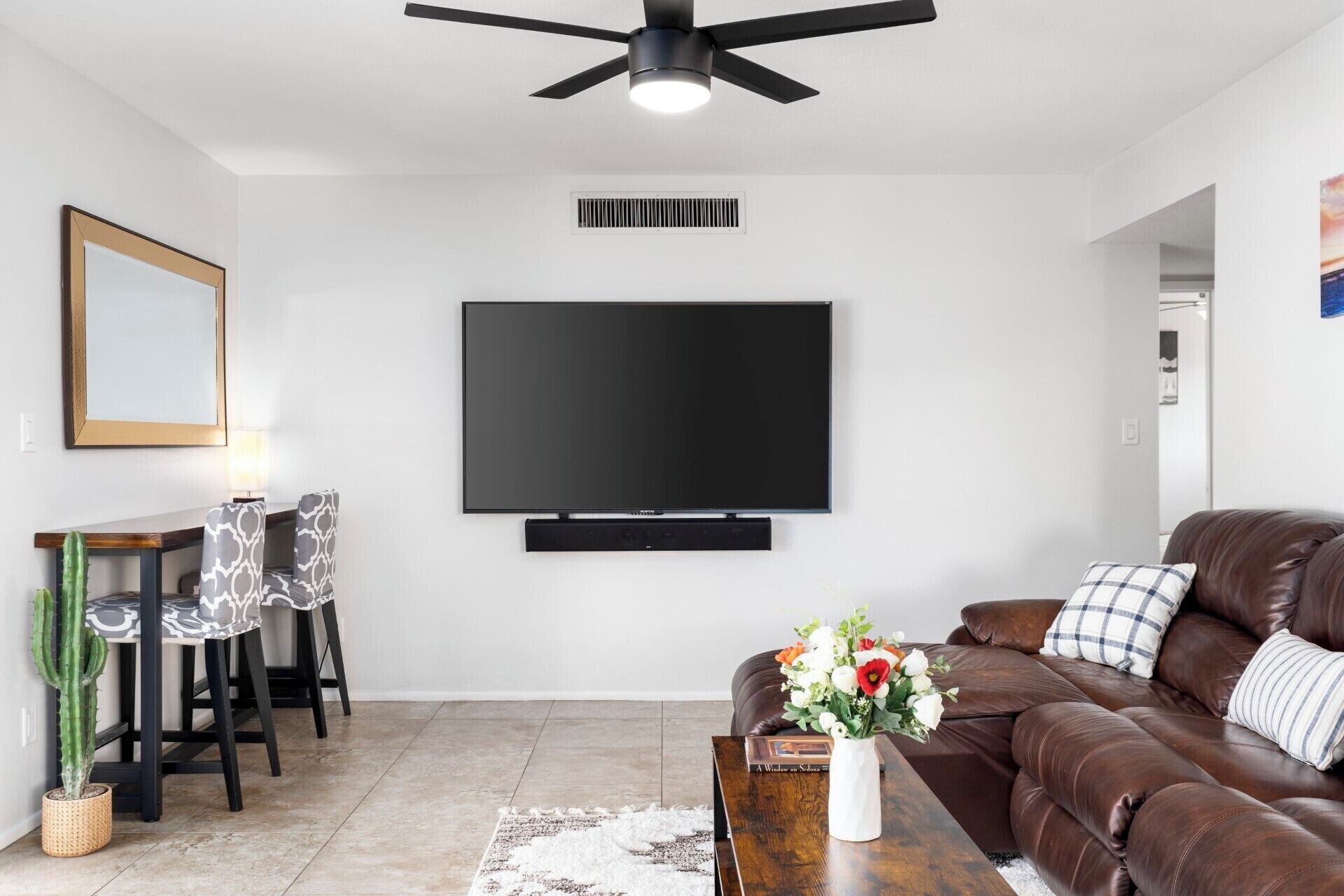 Un salon moderne avec un téléviseur à écran plat fixé au mur, un canapé en cuir marron, un coin repas agrémenté d’un cactus et un ventilateur de plafond.