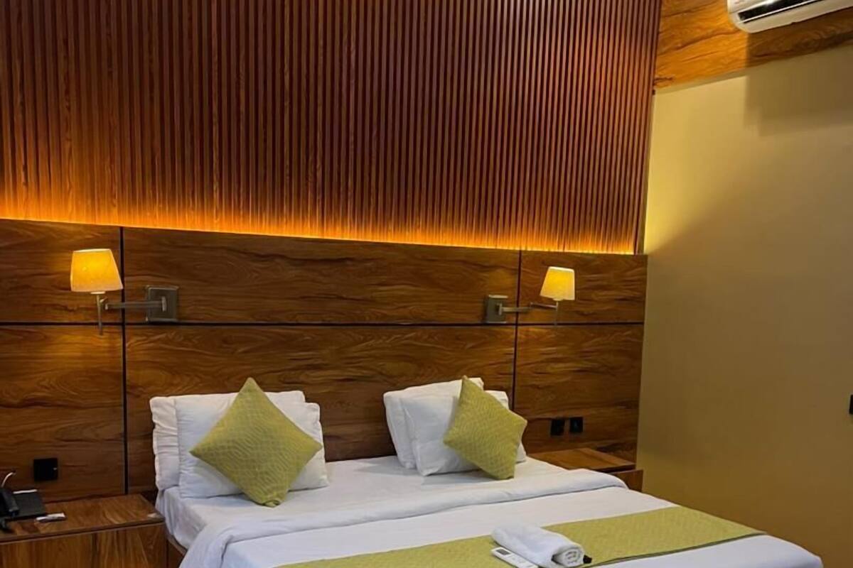 A hotel room with a large bed, two bedside tables, a wooden headboard, and a wall-mounted air conditioner.