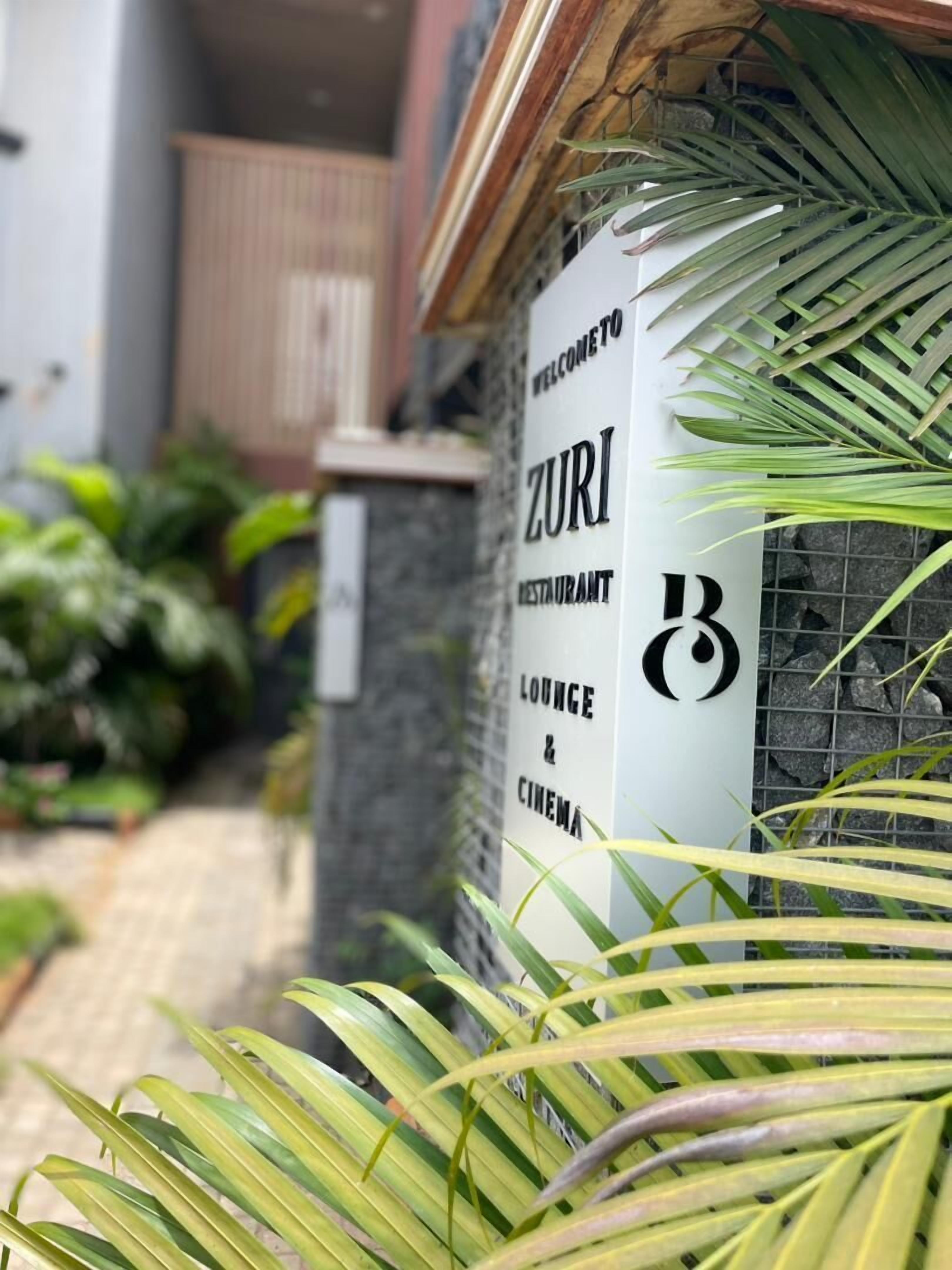 A sign for ZURI Restaurant, Lounge & Cinema, with a stone wall and plants in the background.