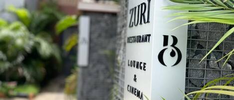 A sign for ZURI Restaurant, Lounge & Cinema, with a stone wall and plants in the background.