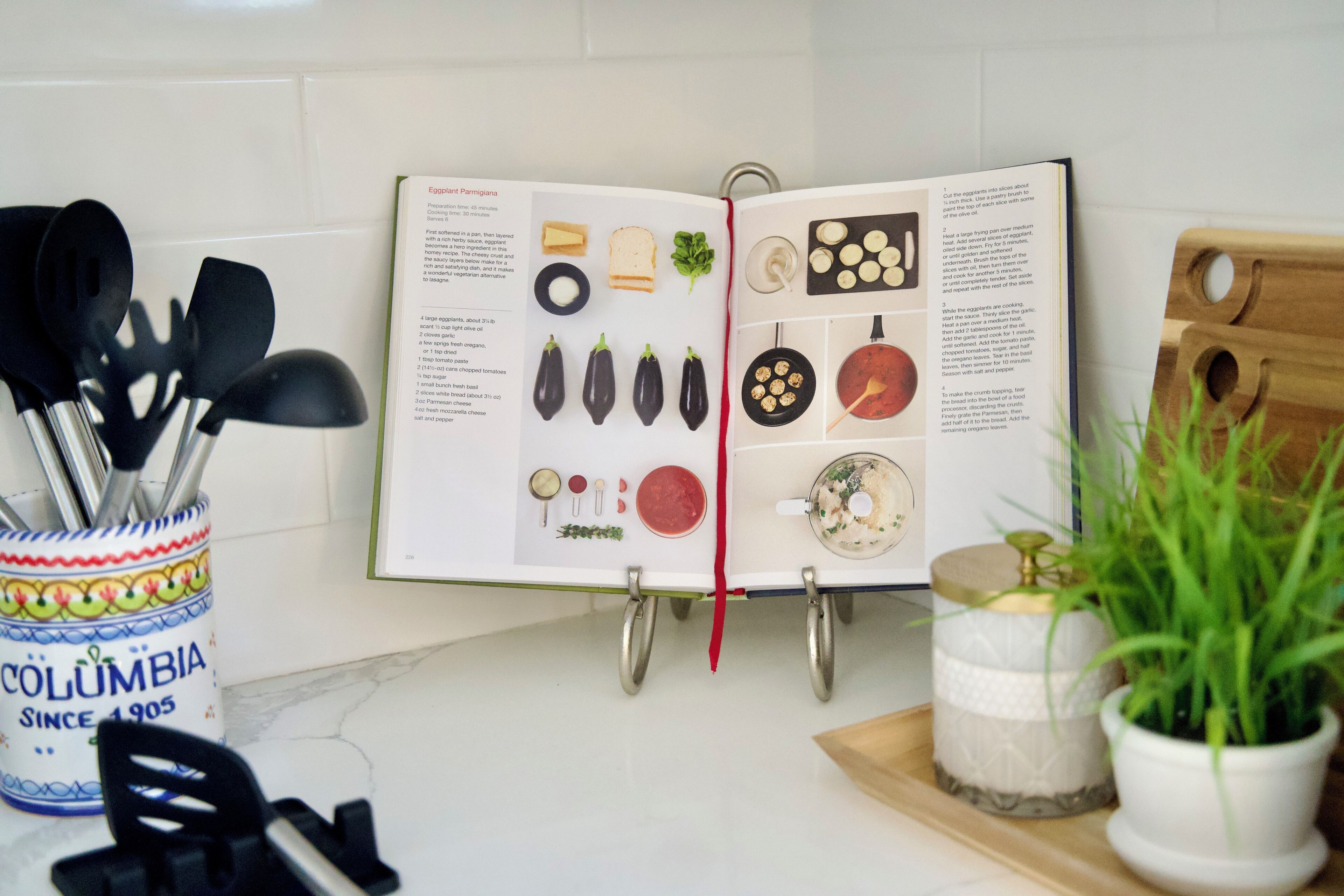 A cookbook open on a kitchen counter with various utensils and a potted plant.