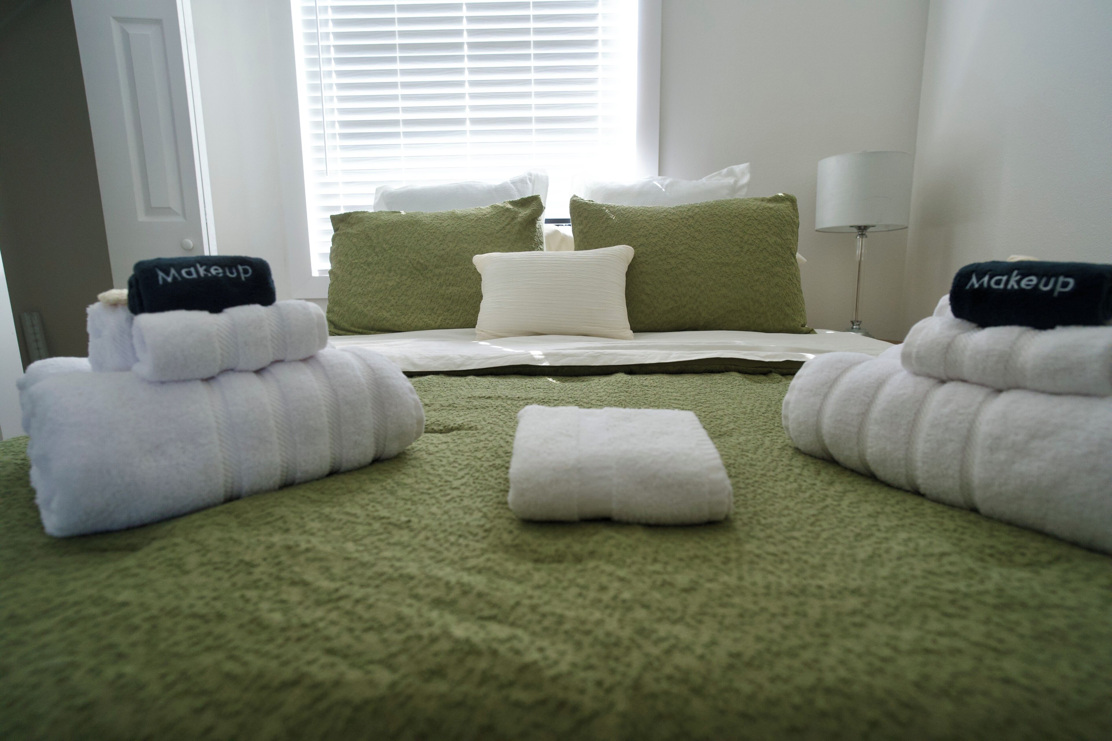 Una cama bien hecha con almohadas verdes y blancas, una manta blanca y dos toallas con la palabra 'Makeup'.