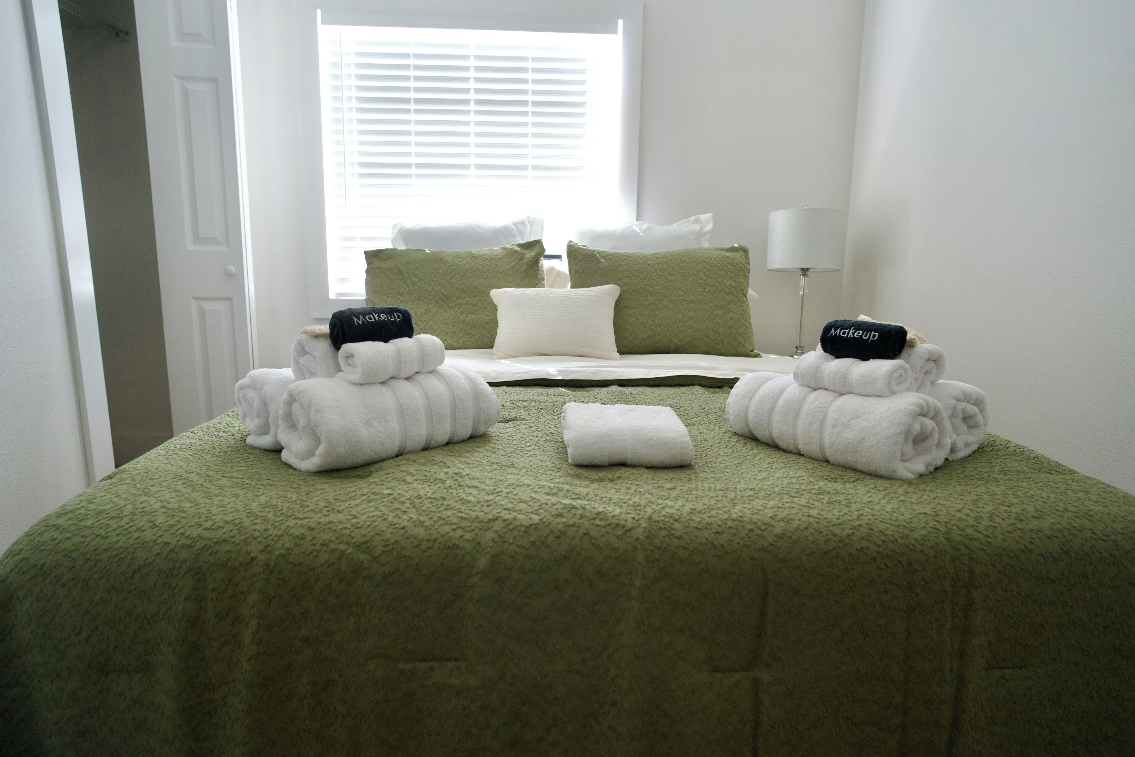 Een net opgemaakt bed met groene beddengoed, witte kussens en handdoeken, inclusief handdoeken met 'makeup'-opdruk.