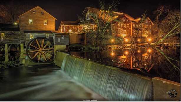 Eine historische Mühle mit einem großen Wasserrad, ein Holzgebäude und ein Restaurant mit beleuchteter Beschilderung.