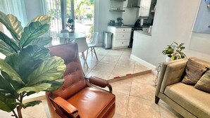 A modern living room with a brown leather sofa, a beige sofa, and a large green plant. There is a dining area with a white table and chairs, and a kitchen in the background. - The Villa unit - The Agustin Guesthouse for Men, a beautiful & sexy resort oasis (Fort Lauderdale)