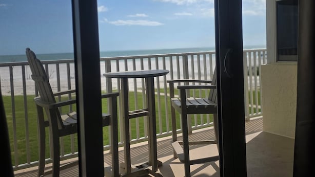 A balcony with two chairs and a small table, overlooking a beach with a clear sky. - Beachfront w Stunning Views Modern Bright (Fort Myers Beach)