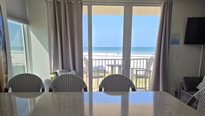 A room with a large window overlooking a beach, a kitchen area with a granite countertop, and wicker furniture. - Beachfront w Stunning Views Modern Bright (Fort Myers Beach)