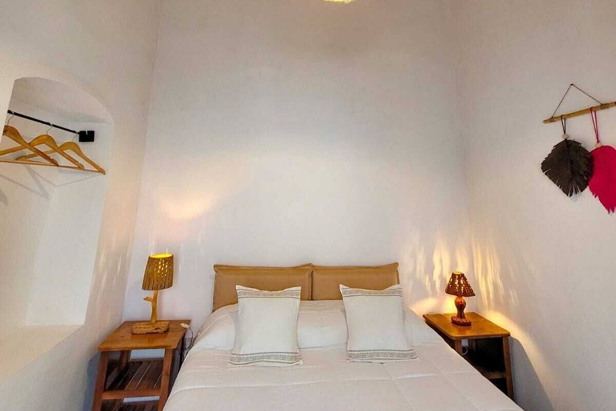 Un dormitorio con techo de madera, una cama con ropa blanca, una lámpara colgante y una mesita de noche pequeña con lámpara.