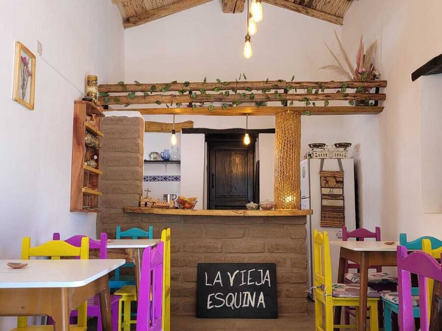 Un intérieur rustique avec des poutres en bois, un panneau noir sur lequel on peut lire « LA YIEJA ESQUINA », et des chaises aux couleurs vives.
