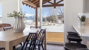 Un comedor con mesa y sillas de madera, un jarrón grande con flores y vistas a montañas nevadas a través de un ventanal.