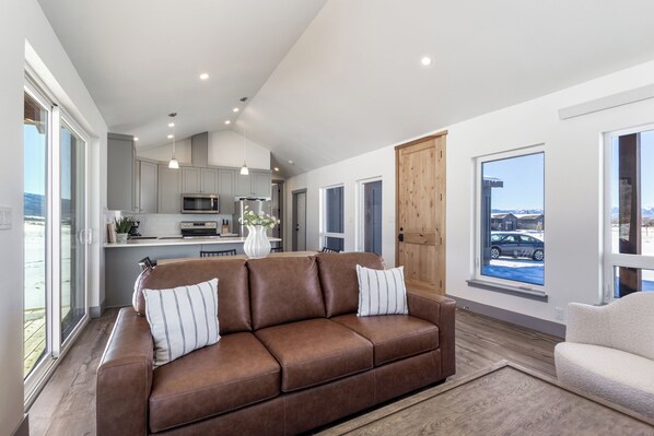 Un salón moderno con un sofá de cuero marrón, un sillón blanco y una cocina al fondo.