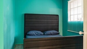 Una habitación con paredes azul verdoso, una cama de madera oscura con dos almohadas, y una ventana con persianas.