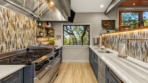 Une cuisine moderne dotée d’un grand îlot central, d’appareils électroménagers en acier inoxydable et offrant une vue sur les arbres par une fenêtre.