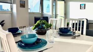 Uma área de refeições com uma mesa posta para dois, com tigelas e pratos azuis, uma pequena planta em vaso e uma televisão ao fundo.