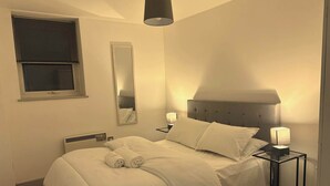 Uma cama bem arrumada com lençóis brancos, uma mesa de cabeceira com abajur, um espelho e uma janela com persianas.