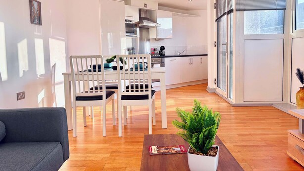 Uma sala de estar moderna com um sofá, uma área de refeições com cadeiras, uma cozinha ao fundo e uma grande janela com persianas.
