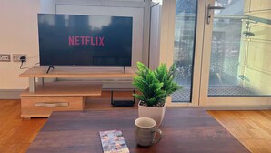 Uma sala de estar moderna com uma grande televisão de tela plana exibindo "NETFLIX", uma mesa de centro de madeira com uma planta e uma caneca, e vista de um edifício por grandes janelas.