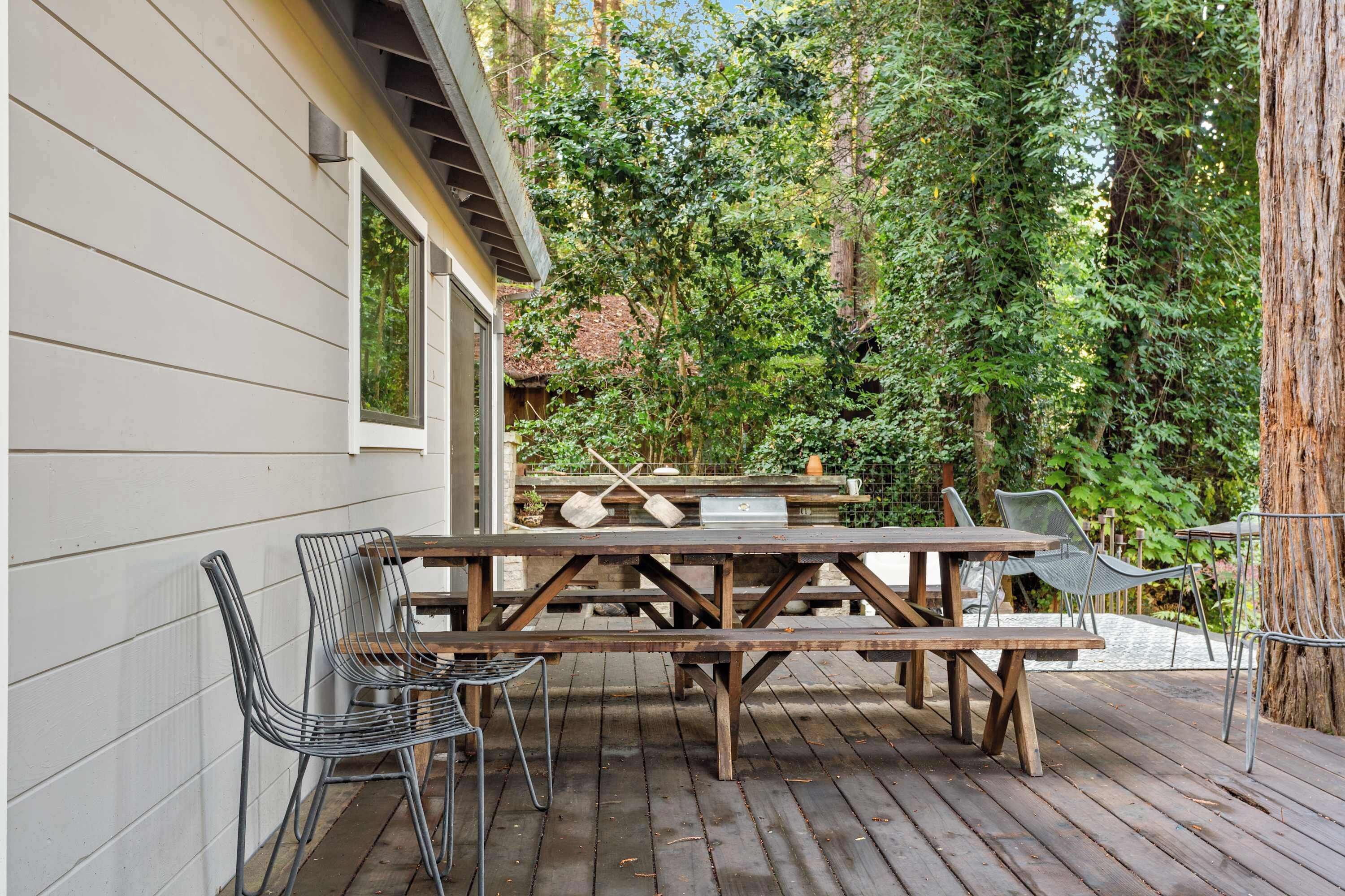 An outdoor patio with wooden picnic tables and metal chairs, surrounded by trees.