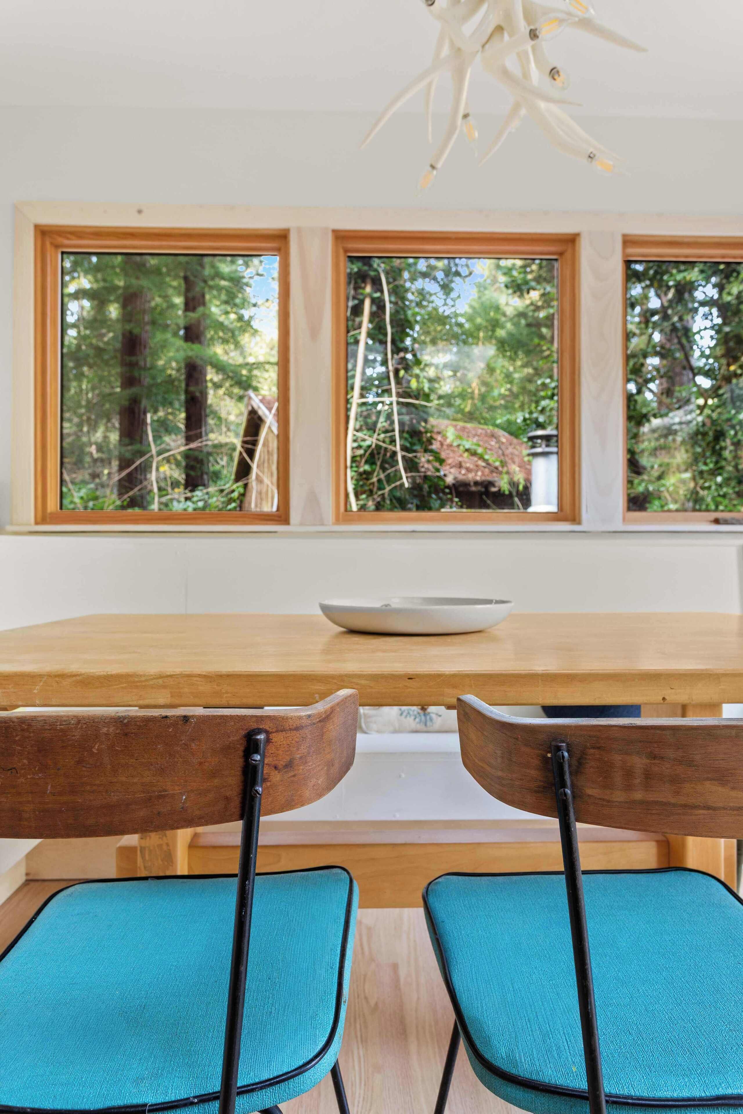 A dining area with a wooden table, two chairs, and a view of a forest through large windows.